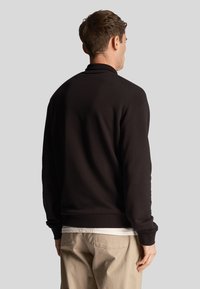 Schwarzer Pullover mit hohem Kragen, gerippten Bündchen und Saum; aus weichem Stoff; von hinten mit einem schlichten weißen Hemd darunter zu sehen.