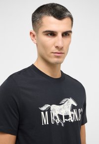 Černé bavlněné tričko s výraznou grafikou bílého koně a nápisem "MUSTANGS" v elegantním písmu. Klasický kulatý výstřih.