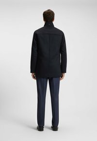 Mann, der mit dem Rücken zu sehen ist, trägt einen schwarzen Mantel, navyfarbene Anzughose und schwarze Schuhe vor einem schlichten hellgrauen Hintergrund.