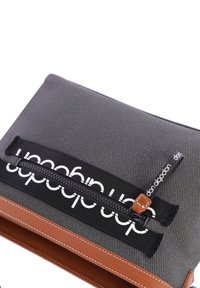 Bolsa de tela negra con una superficie texturizada, detalles en cuero marrón, apertura con cremallera y una marca en blanco en negrita en la parte frontal.
