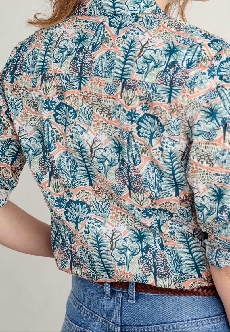 Femme portant une chemise colorée avec un motif botanique abstrait en bleu, vert et orange, associée à un jean bleu et une ceinture tressée marron.