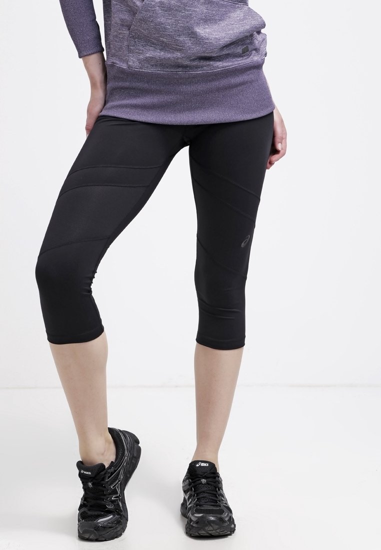 Personne portant des leggings de sport noirs coupés, des chaussures de course noires et un haut à manches longues violet, debout les jambes légèrement écartées.
