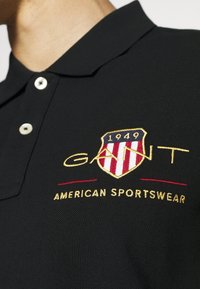 Svart polo-shirt med krage och tre knappar. Broderad sköld med röd och vit rand, guldtext "GANT" och "AMERICAN SPORTSWEAR."