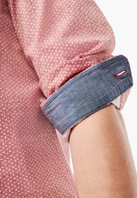Camisa de cuadros rojos y blancos con puños de denim azul remangados y costuras rojas en contraste a lo largo de la manga, mostrando la textura de la tela.