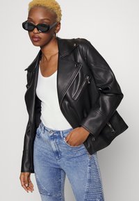 Jaqueta de motociclista em couro preto com fechos prateados, usada sobre uma t-shirt branca, combinada com jeans claros e desgastados. Óculos de sol pretos.