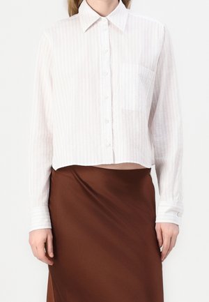 Vrouw draagt een cropped wit overhemd met subtiele strepen en een borstzak, gecombineerd met een hooggetailleerde bruine rok.