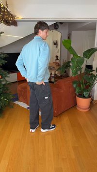 Νεαρός άνδρας με ανοιχτό μπλε μπουφάν και μαύρο παντελόνι στέκεται σε ένα σαλόνι με χριστουγεννιάτικο δέντρο, πορτοκαλί καναπέ και μεγάλο φυτεμένο φυτό.