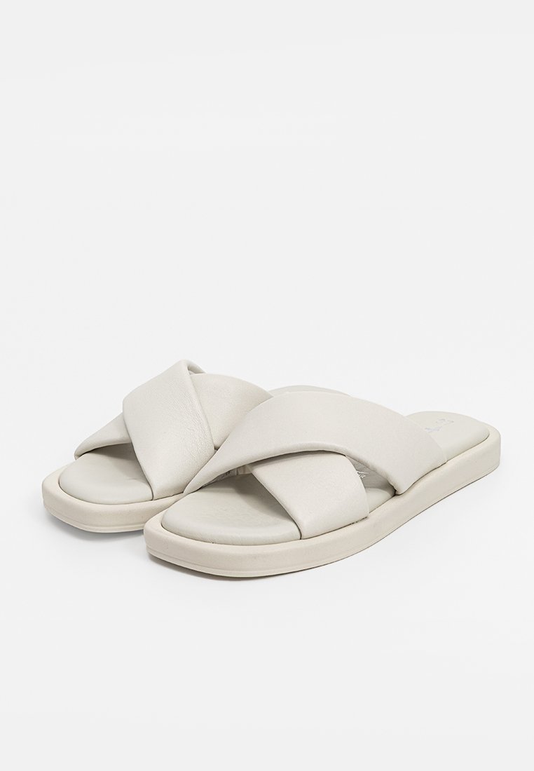 Cremfarbene Rutschschuhe mit dicken, überkreuzten gepolsterten Riemen und flachen, gepolsterten Sohlen auf einem weißen Hintergrund.