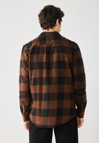 Camisa de franela a cuadros marrón y negra con mangas largas, yugo en la parte posterior y puños abotonados. Textura suave y ajuste casual.