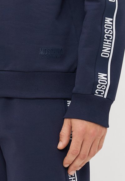 Marineblå Moschino joggedress med merkevarebånd på ermene og buksene, hånden hviler ved bukselommen.