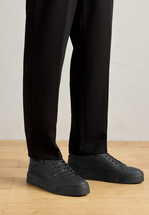 Crne tenisice od sintetičkog materijala, s ravnim potplatima, okruglim nožnim prstima i dizajnom sa vezanjem, uz detalj bočnog patentnog zatvarača.