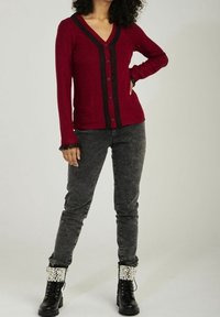 Czerwony kardigan z dekoltem w serek, czarnym zapięciem na guziki i koronkowym wykończeniem rękawów, w połączeniu z ciemnoszarymi dżinsami i czarnymi sznurowanymi botkami z perłowymi akcentami.