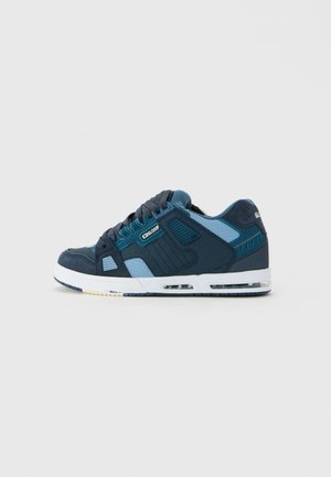 Sneaker de tip low-top de culoare albastră închisă și albastră deschisă, cu șireturi negre și perna de aer vizibilă în talpa albă, văzut de pe lateral pe un fundal simplu.