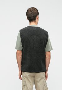 Juodas pūkinis liemenė su tekstūruotu paviršiumi, be rankovių dizainu ir laisvu siluetu, užsidėta ant šviesiai žalios marškinėlių ir smėlio spalvos kargo kelnių.