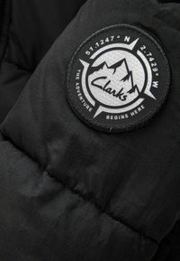 Svart puffig tyg med en cirkulär remsa. Remsan har vita berg, texten "Clarks" och koordinater. Sömmen syns på materialet.