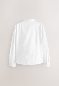 Blouse blanche à manches longues avec col festonné et poignets boutonnés, vue de dos sur un fond neutre uni.