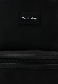 Zaino nero con tessuto testurizzato, chiusura con zip e una patch rettangolare del logo Calvin Klein nera con scritta bianca.