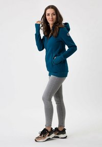 Felpa con zip color teal, dotata di tasche laterali, tessuto elasticizzato e design aderente. Abbinata a leggings grigi chiari e scarpe sportive multicolori.