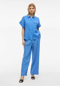 Completo di camicia e pantaloni in tessuto testurizzato. Maniche corte, chiusura con bottoni sul davanti e tasca sulla camicia; pantaloni a gamba larga. Sandali neri con tacco.