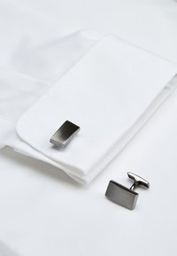 Srebrni manžetni prikazani pored manžete bijele košulje. Manžetni imaju minimalistički pravokutni dizajn s četkanom teksturom.