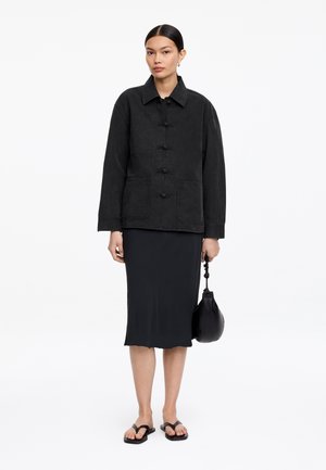 Mujer vestida con una chaqueta negra abotonada, falda midi negra, sandalias negras, sosteniendo un pequeño bolso negro con una correa anudada.