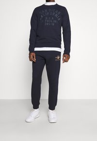Navy Sweatshirt mit "SAN DIEGO CALIFORNIA" in dunkler Schrift, kombiniert mit navyfarbenen Sweatpants mit einem goldenen Logo, getragen mit weißen Sneakers.