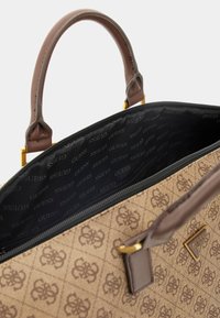 Sac à main à motif tan avec des poignées en cuir brun foncé et des accessoires en doré. L'intérieur doublé présente le logo "Guess" répété sur un fond noir.