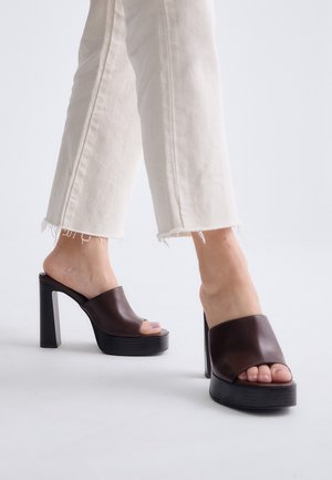 Persona que lleva pantalones cortos de color beige claro y zuecos de plataforma de tacón alto color marrón oscuro con dedos abiertos sobre fondo blanco.