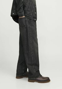 Blugi din denim negri, cu o croială lejeră, prezentând detalii cusute, buzunare laterale și un design cu picioare drepte, asortați cu pantofi maro.