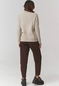 Kobieta stojąca tyłem, ubrana w beżowy sweter z golfem o żeberkowanej fakturze, ciemne brązowe spodnie i czarne sneakersy na platformie, na jednolitym tle.