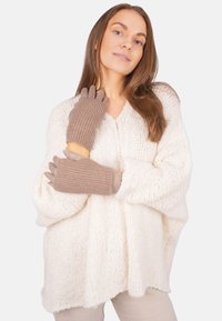 Frau in einem cremefarbenen, grob gestrickten Cardigan und beigen gerippten Handschuhen, die mit einer Hand an ihrem Hals vor einem weißen Hintergrund posiert.