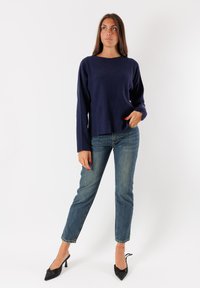 Donna in piedi con jeans, maglione a costine blu navy e scarpe con tacco nere, che guarda leggermente di lato con una mano in tasca.