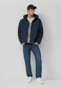 Granatowa kurtka puchowa z kapturem, zapinana na zamek, z bocznymi kieszeniami; noszona na jasnym swetrze i w połączeniu z niebieskimi dżinsami oraz białymi trampkami.