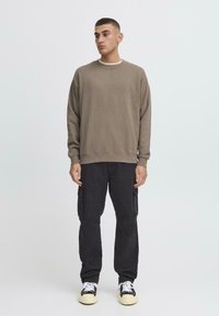 Brun crewneck genser, baggy svarte cargo-bukser med sidelommer, hvite joggesko, enkel bakgrunn, nøytrale farger, avslappet passform.