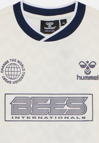 Maillot de sport blanc avec col bleu marine, logo "hummel", globe avec le texte "Change the world through sport" et branding "BEES Internationals".