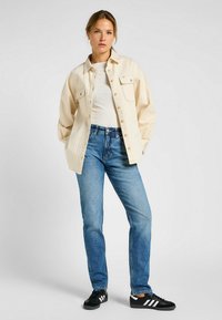 Camisa beige de gran tamaño con bolsillos frontales, combinada con jeans rectos de color azul claro y zapatillas negras con franjas blancas.