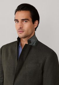 Mann mit dunklem Haar trägt eine dunkle Jacke mit hohem Kragen über einem blauen Hemd und schaut leicht zur Seite vor schlichtem Hintergrund.