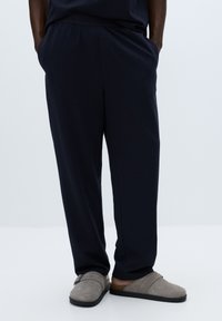 Voľné nohavice námorníckej modrej farby vyrobené z mäkkej tkaniny, s bočnými vreckami a hladkou textúrou, doplnené o sivú obuv na naskočenie.