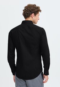 Uomo con capelli ricci che indossa una camicia nera a maniche lunghe e pantaloni grigi, girato per mostrare la schiena della camicia e parte del suo profilo.