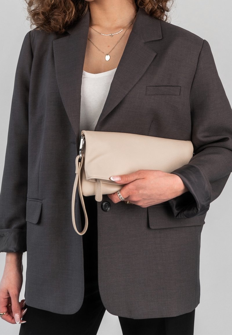 Femme portant un blazer gris foncé et un haut blanc tient une pochette beige avec une fermeture éclair argentée et une sangle sur fond gris clair.