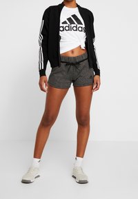 Chaqueta negra con cremallera y rayas blancas, camiseta blanca con logotipo negro, pantalones cortos grises y zapatillas ligeras con calcetines blancos.