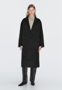 Lungo cappotto di lana nera, vestibilità oversize, rever a becco, due bottoni, dettagli minimi; abbinato a pantaloni grigi texturizzati e scarpe nere.