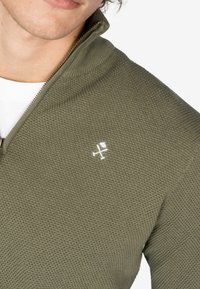 Suéter de cremallera verde oliva con un patrón de punto texturizado y un pequeño logo blanco en el pecho. Cuello alto y puños acanalados.