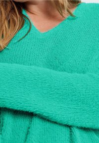 Turkusowy sweter wykonany na drutach, z dekoltem w kształcie V, miękką fakturą i luźnym krojem. Tkanina wydaje się puszysta, podkreślając wygodę i ciepło.