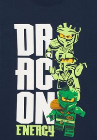 Tamsiai mėlyna marškinė su trimis Lego nindzių personažais žalia ir geltona spalvomis, o centre ryškiai baltais žodžiais parašyta "DRAGON ENERGY".