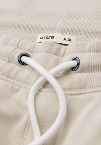 Pantalones con cintura elástica beige y cordón blanco que pasa por ojales de metal negro, etiquetados "ombre" talla M eur 48.