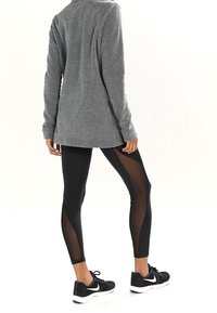 Czarne legginsy z przezroczystymi siateczkowymi wstawkami na zewnętrznych nogawkach, zestawione z szarym długim topem. Czarne buty sportowe dopełniają outfit.