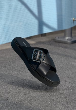 Sort lædersandal med tyk sål og stor sølvspænde, der svæver over en grå asfaltflade udendørs.