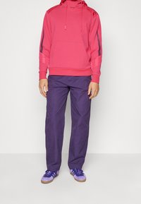 Różowa bluza z kapturem i troczkami, z dużą kieszenią i czarnymi paskami po bokach, w zestawie z fioletowymi spodniami. Buty fioletowe z br ązowymi podeszwami.