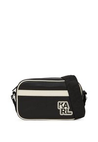 Sac en bandoulière noir avec une texture lisse, présentant des rayures blanches, un patch logo et une sangle réglable. Fermeture zippée en haut.